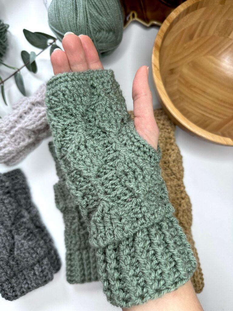 A hand with a crochet green fingerless glove on is palm up over a white table with more gloves on as well as a wooden bowl, greenery and green yarn. The crochet glove features textured leaves of the same colour