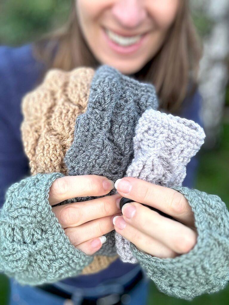 2 hands are holding out 3 different sized and different coloured crochet mittens in front of them, whilst also wearing a pair of green crochet fingerless gloves. The mittens all have the same trailing leaves stitch pattern on them