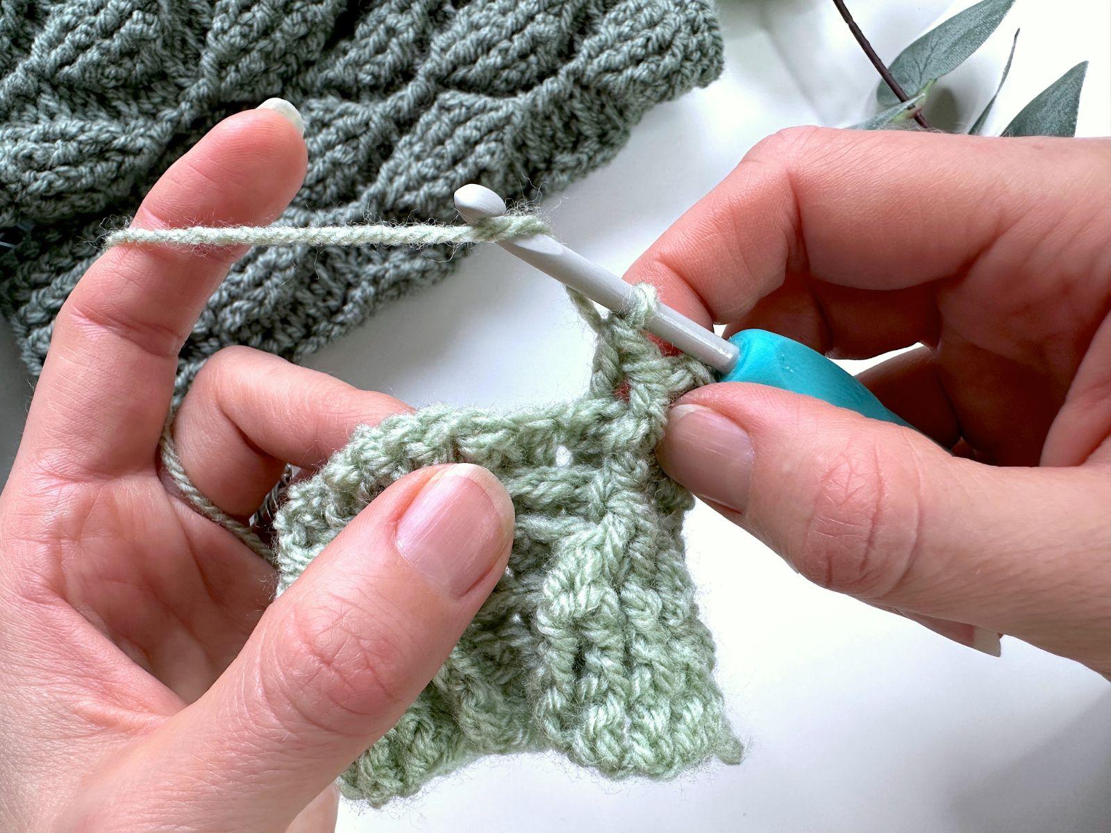 2 hands hold a green crochet sample and are using a crochet hook to wrap the yarn around