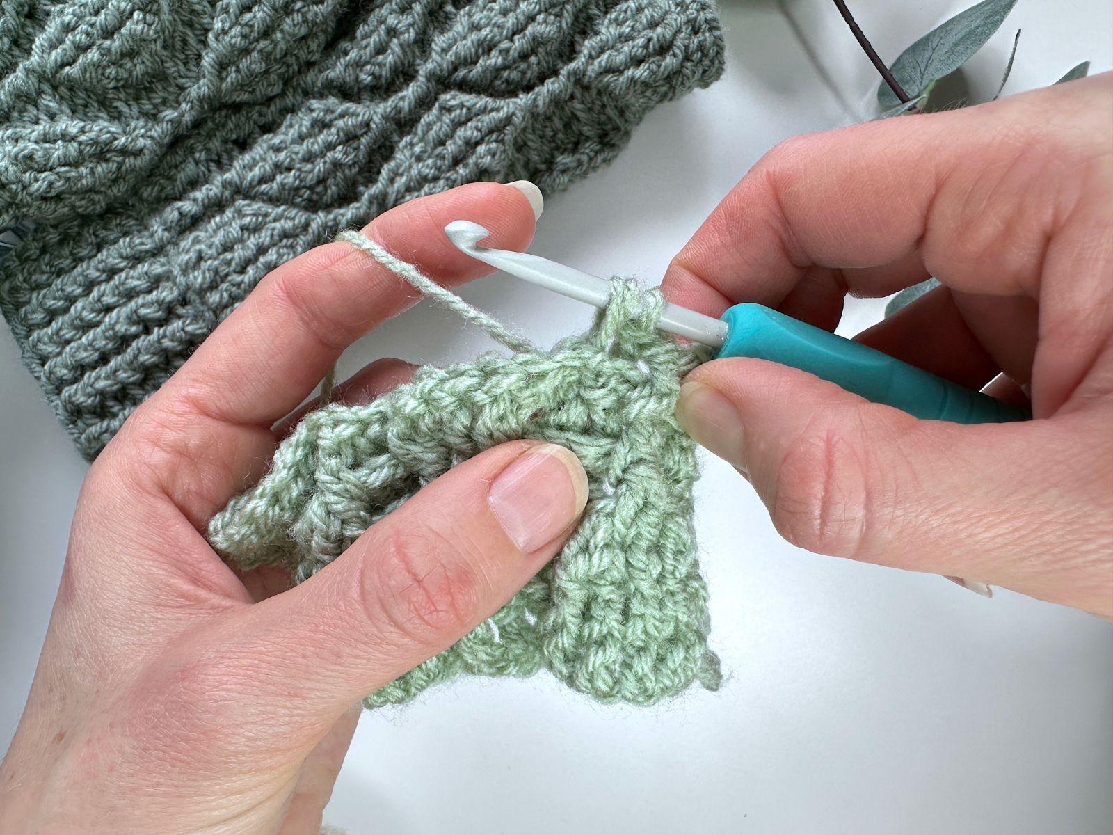 2 hands are partway through making a crochet stitch with green yarn and a turquoise hook