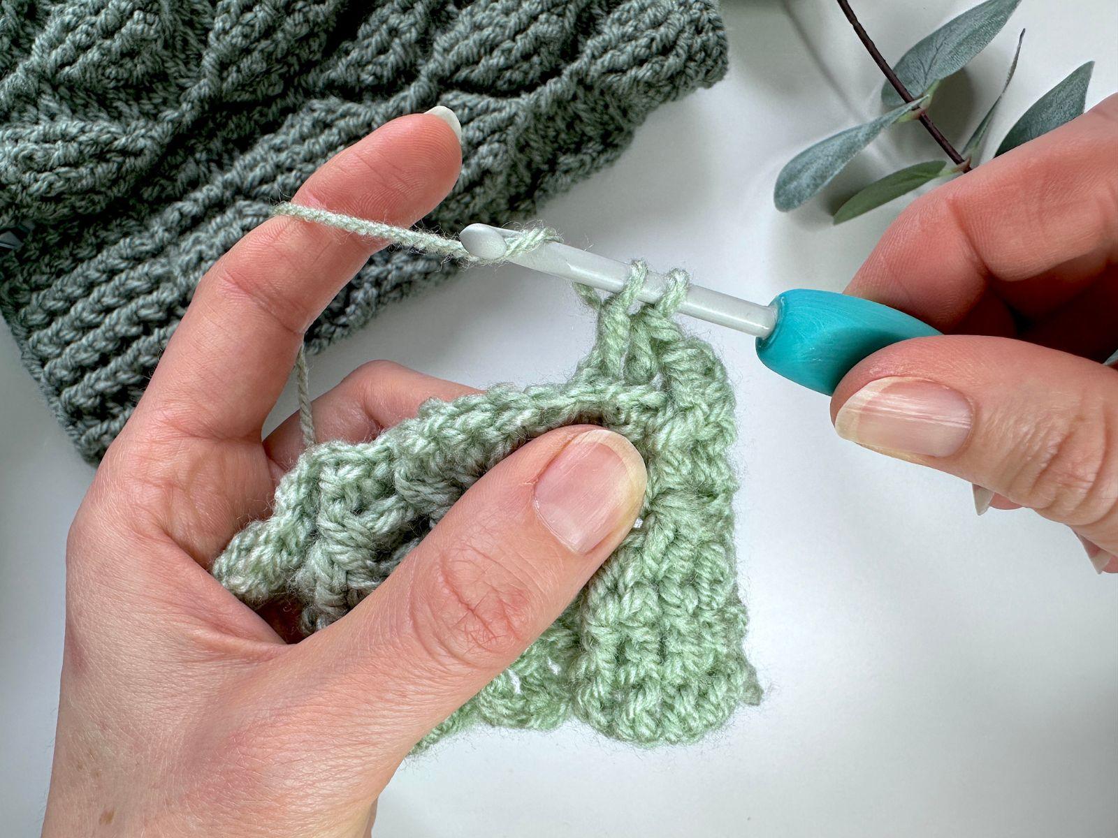A crochet hook and 2 hands wrap yarn around the hook to make a crochet stitch
