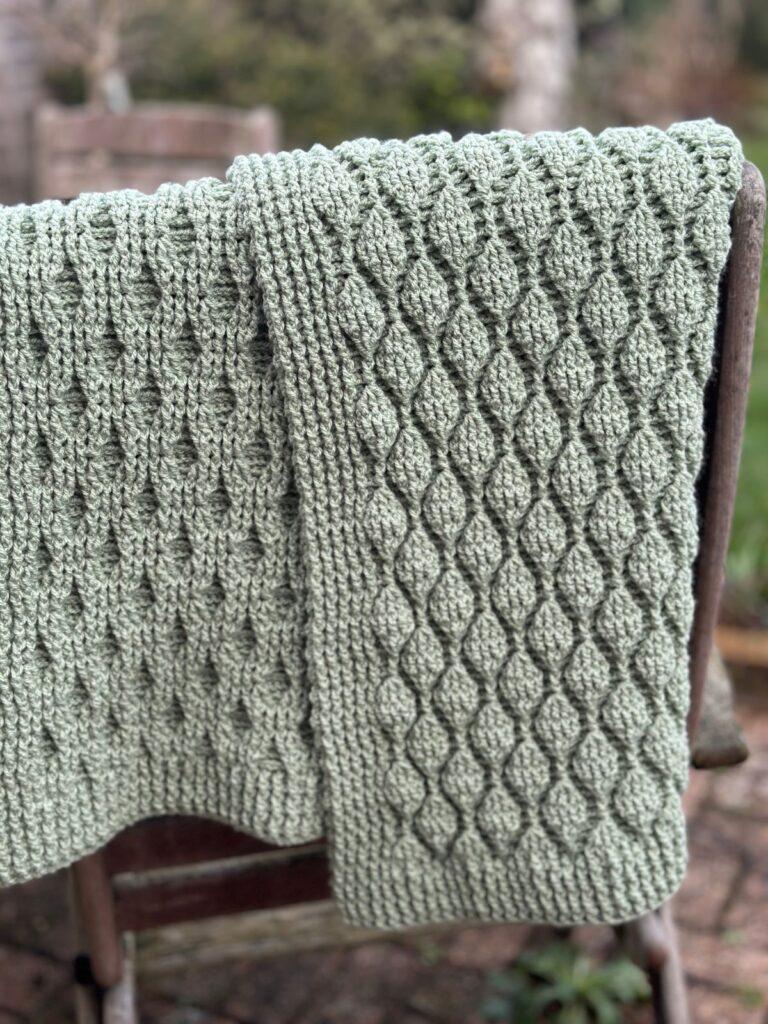 A green textured crochet blanket is folded and hung over a chair so that the front and back sides are showing