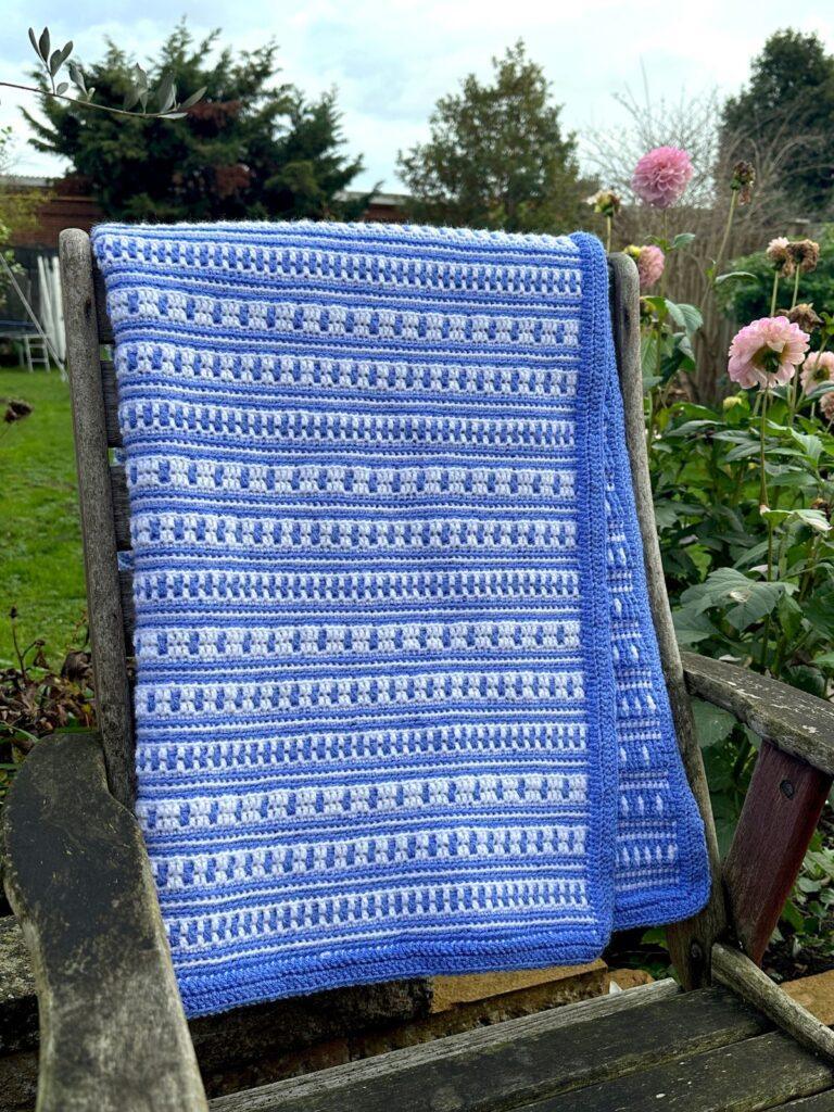 A blue and white striped crochet blanket hangs on the back of a chair in a garden