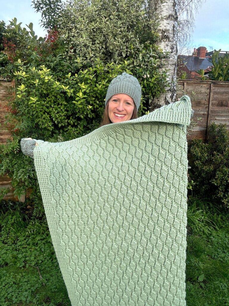 A lady stands in a garden wearing a green crochet hat and holding up a textured green crochet blanket