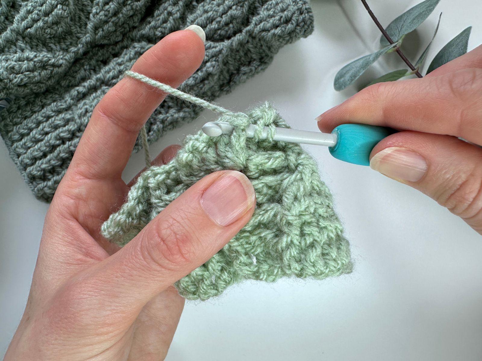 A crocheter shows a sample of green fabric and has inserted a crochet hook into the fabric to make a post stitch