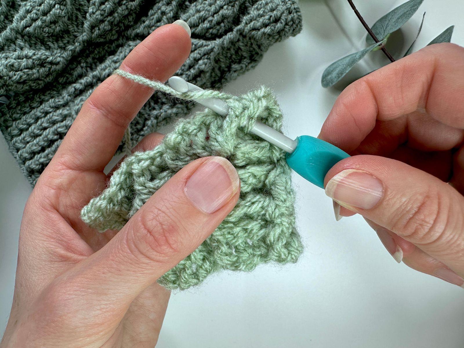 2 hands are holding a crochet hook and crochet sample and are making a stitch