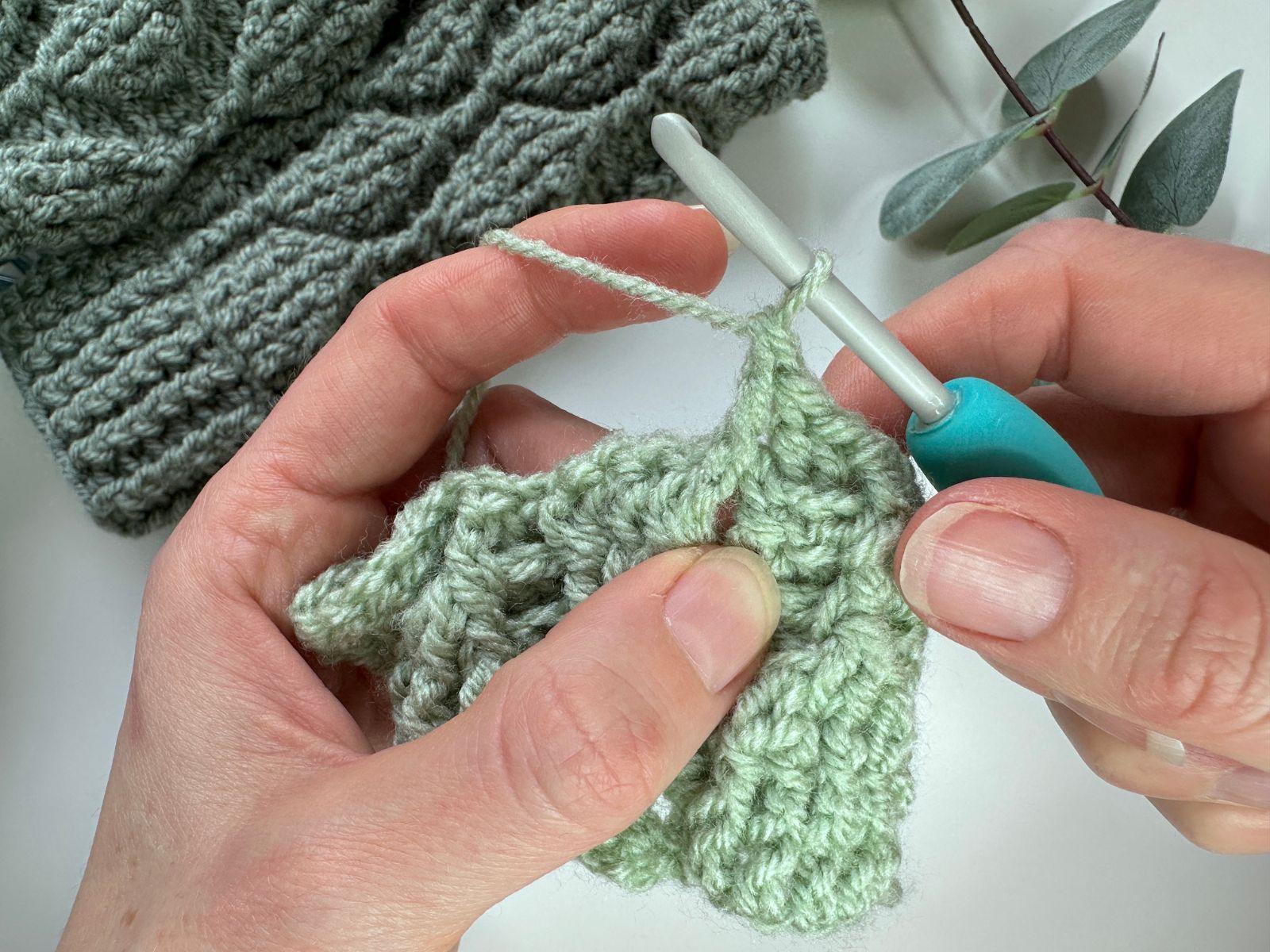 A completed front post double crochet stitch in green yarn is held by 2 hands and there is also a hook