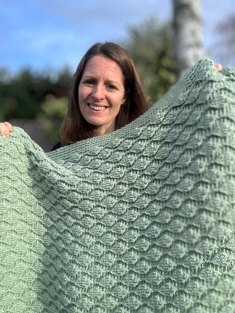 A smiling lady holds up a green crochet blanket with leaves trailing over it in texture