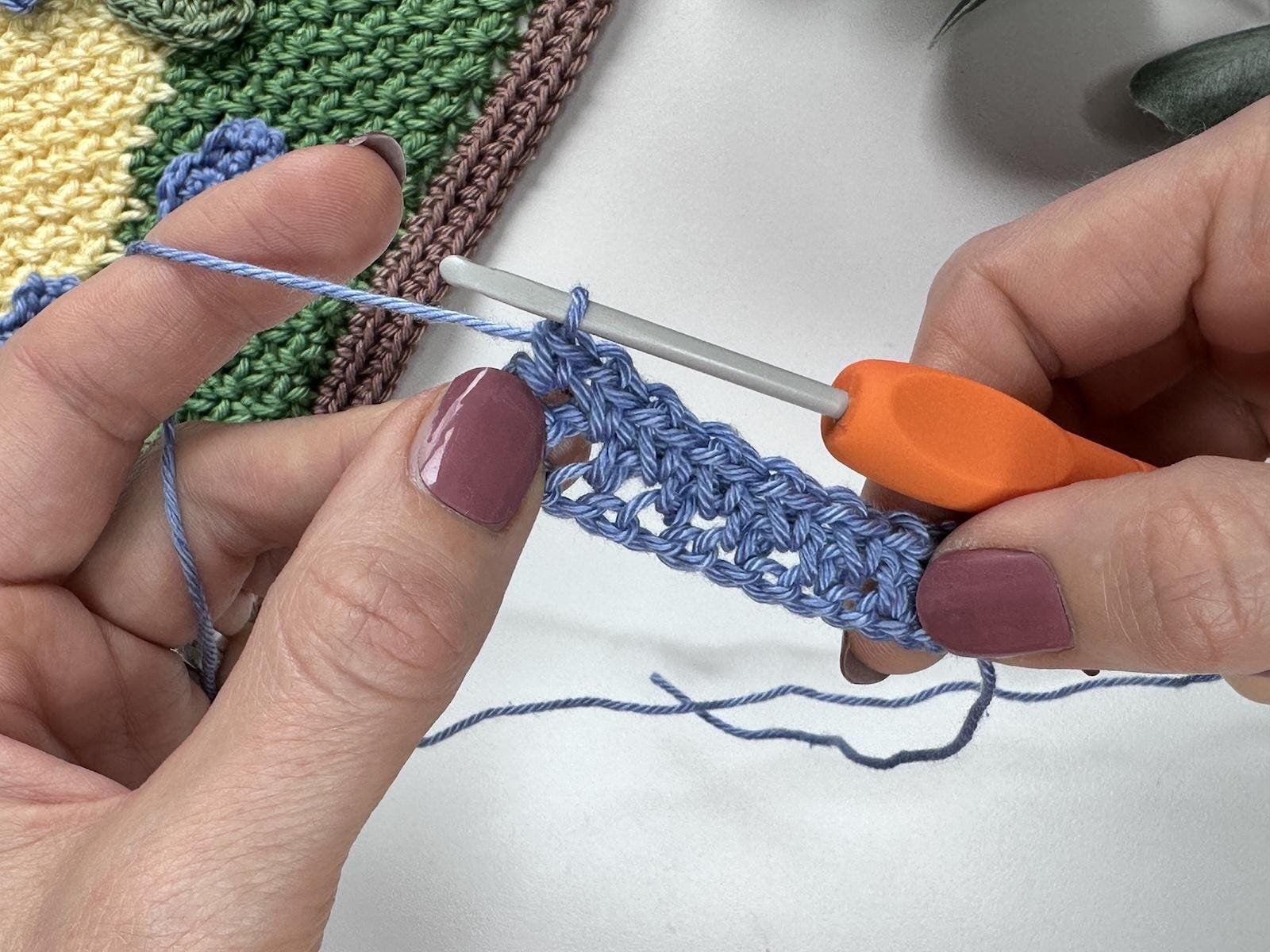 A small completed sample of crochet moss stitch in blue yarn held in 2 hands with an orange crochet hook