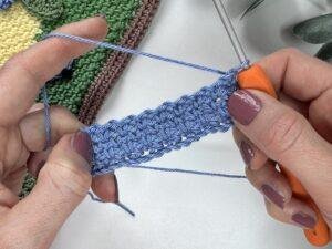 2 hands hold a blue sample of the crochet moss stitch