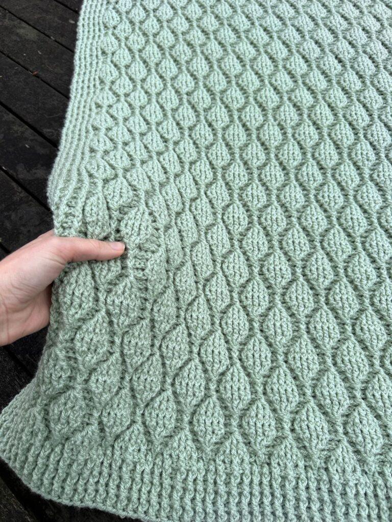A textured green crochet blanket with leaves running up it in post stitches. A hand holds the edge of the blanket
