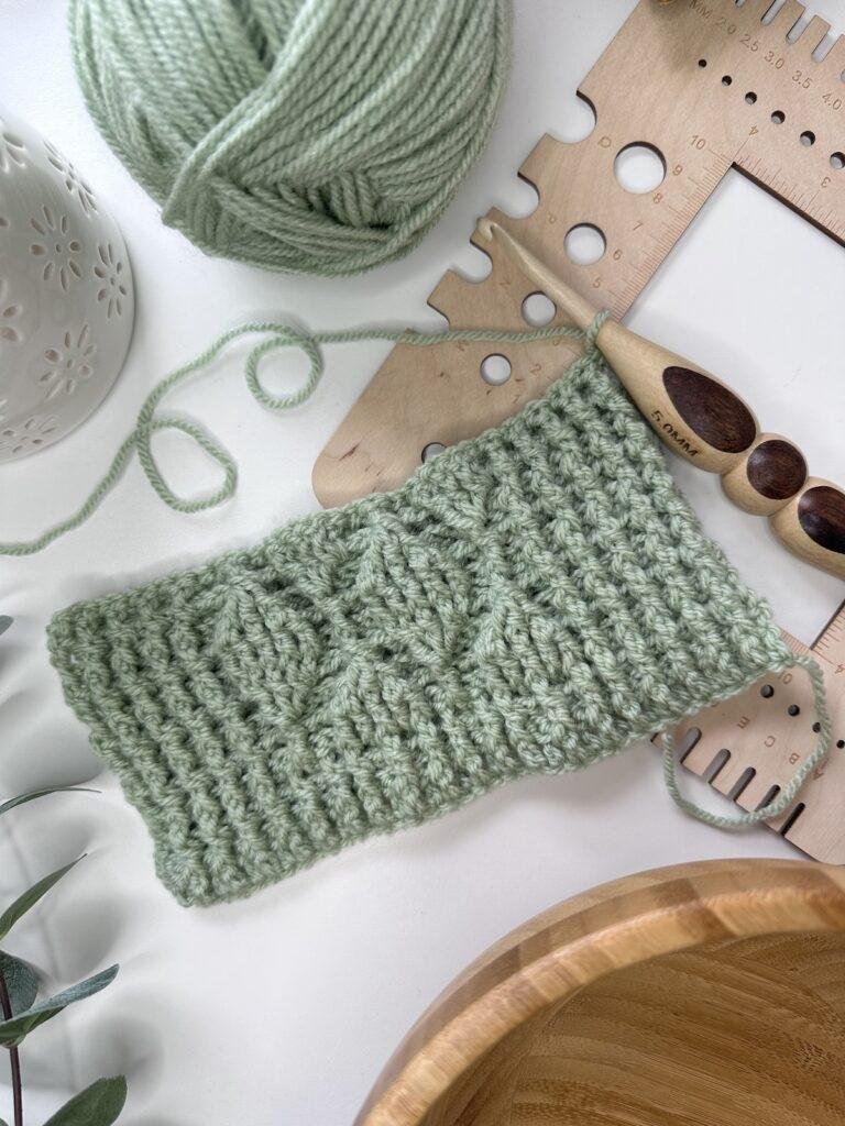 a flatlay of a green crochet textured sample with a wooden hook, gauge tool and yarn around