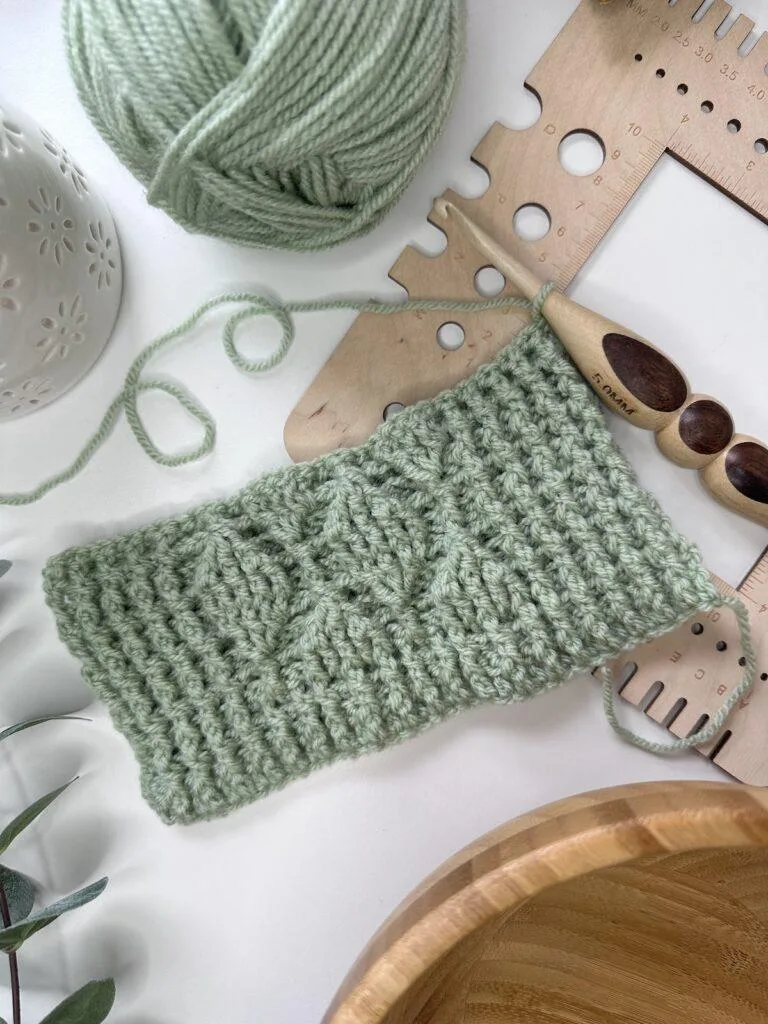 a flatlay of a green crochet textured sample with a wooden hook, gauge tool and yarn around