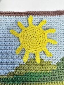 A crochet sunshine is laid on a crochet background which looks like hills and sky in tapestry crochet
