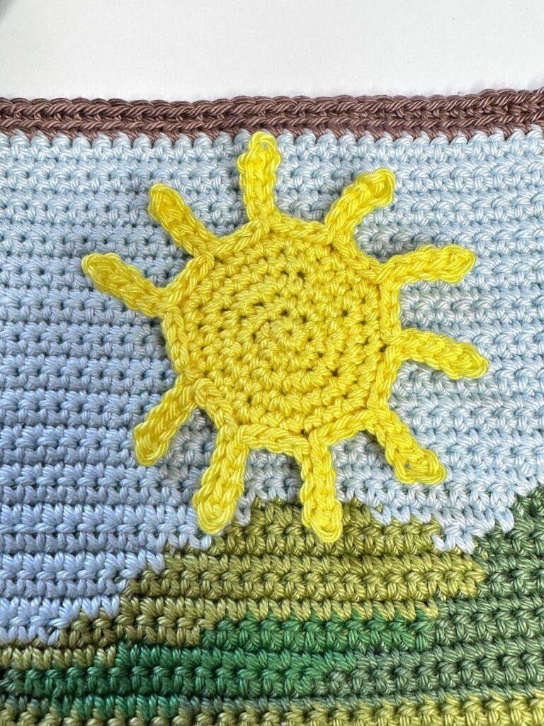 A crochet sunshine is laid on a crochet background which looks like hills and sky in tapestry crochet
