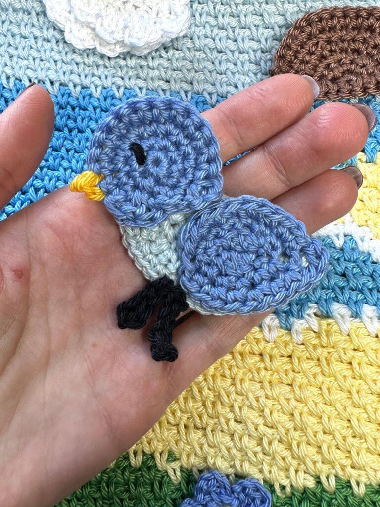 A hand holds a crochet bird applique above a sea background
