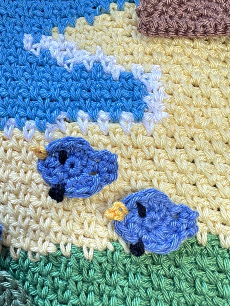 2 tiny crochet blue chicks are placed on a crochet seaside landscape