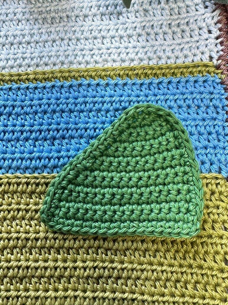 A green crochet hill shape is sewn onto a green and blue crochet background