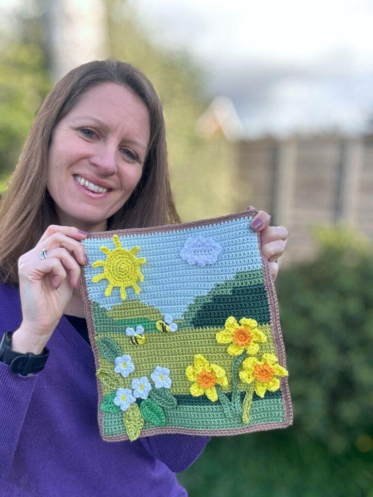 A lady holds up a spring crochet scene with sun, daffodils, small flowers and hills