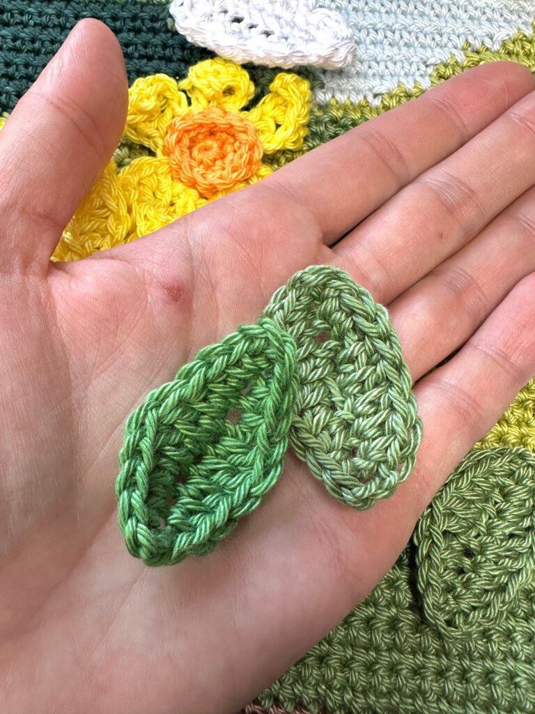 a hand holds 2 small crochet leaves with a crochet landscape underneath