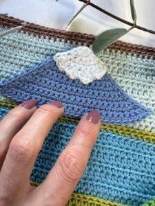 Fingers touch a crochet applique mountain holding it on a blue striped crochet background