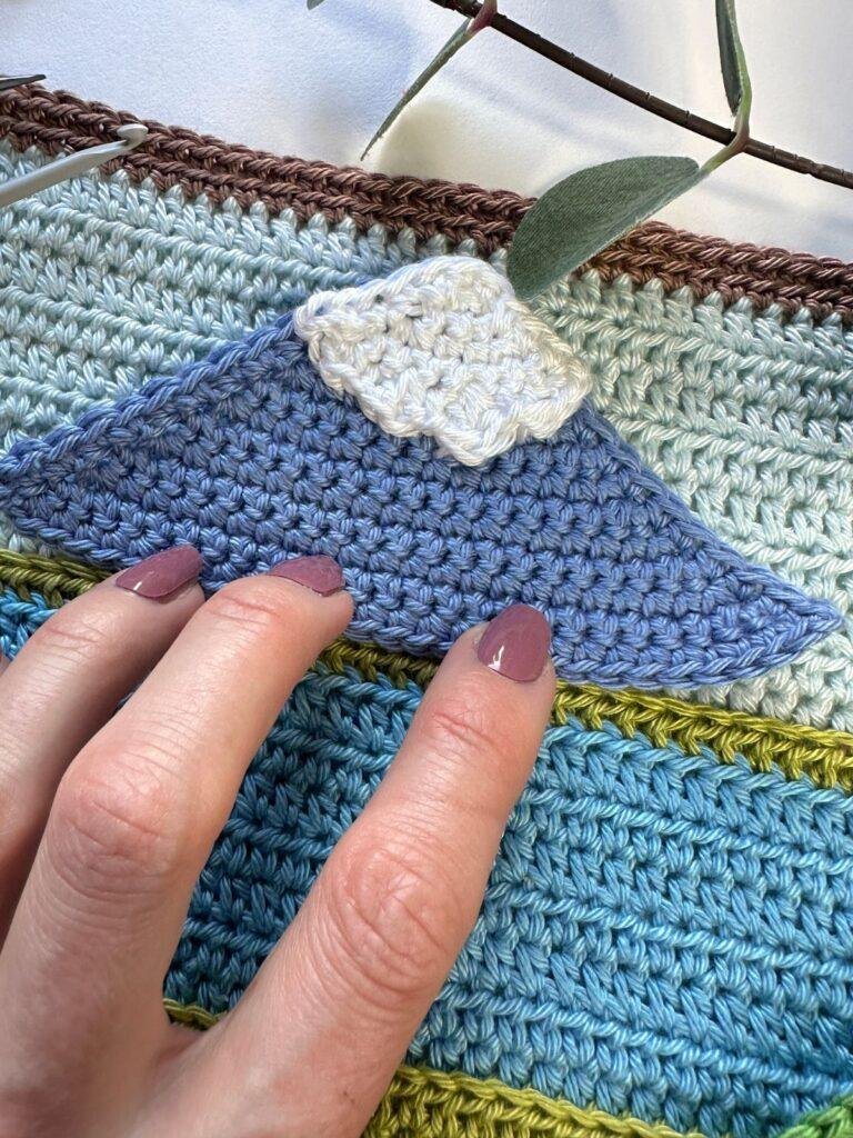 Fingers touch a crochet applique mountain holding it on a blue striped crochet background