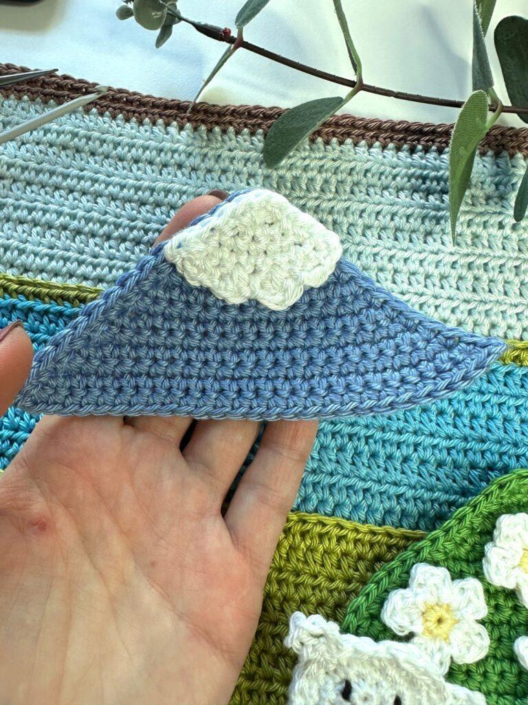 a hand holds up a flat crochet mountain above a blue and green crochet background
