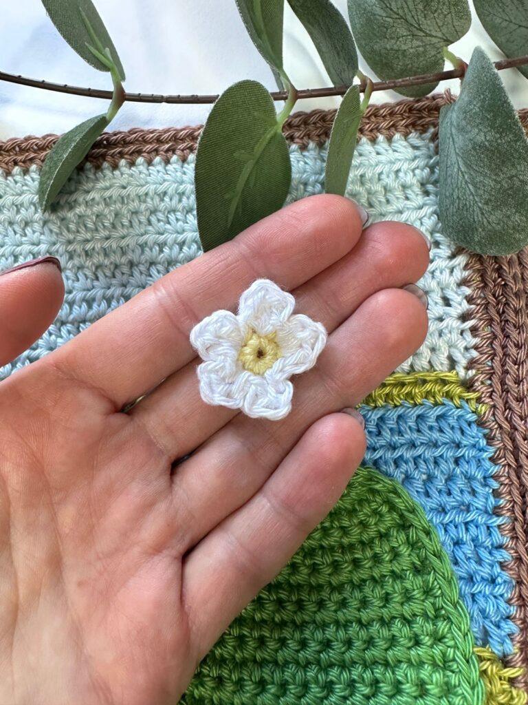 a crochet pansy is held in a hand on top of a crochet scenery