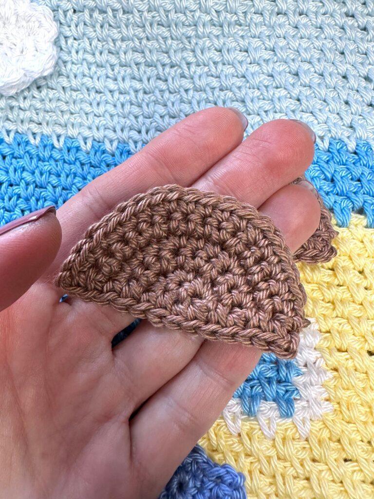 A hand a holding a brown semi circle shaped crochet rock in front of a blue and yellow crochet background