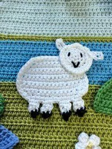 a white crochet sheep applique with a cute face on a green and blue crochet background