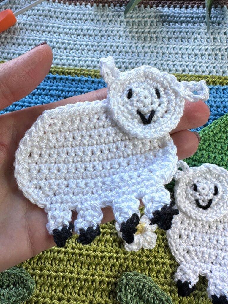 A hand holds a white crochet sheep applique with another in the background