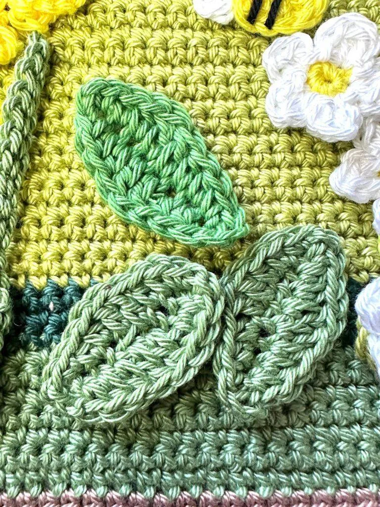 3 green crochet leaves are sewn onto a green crochet background with flowers in the top corner