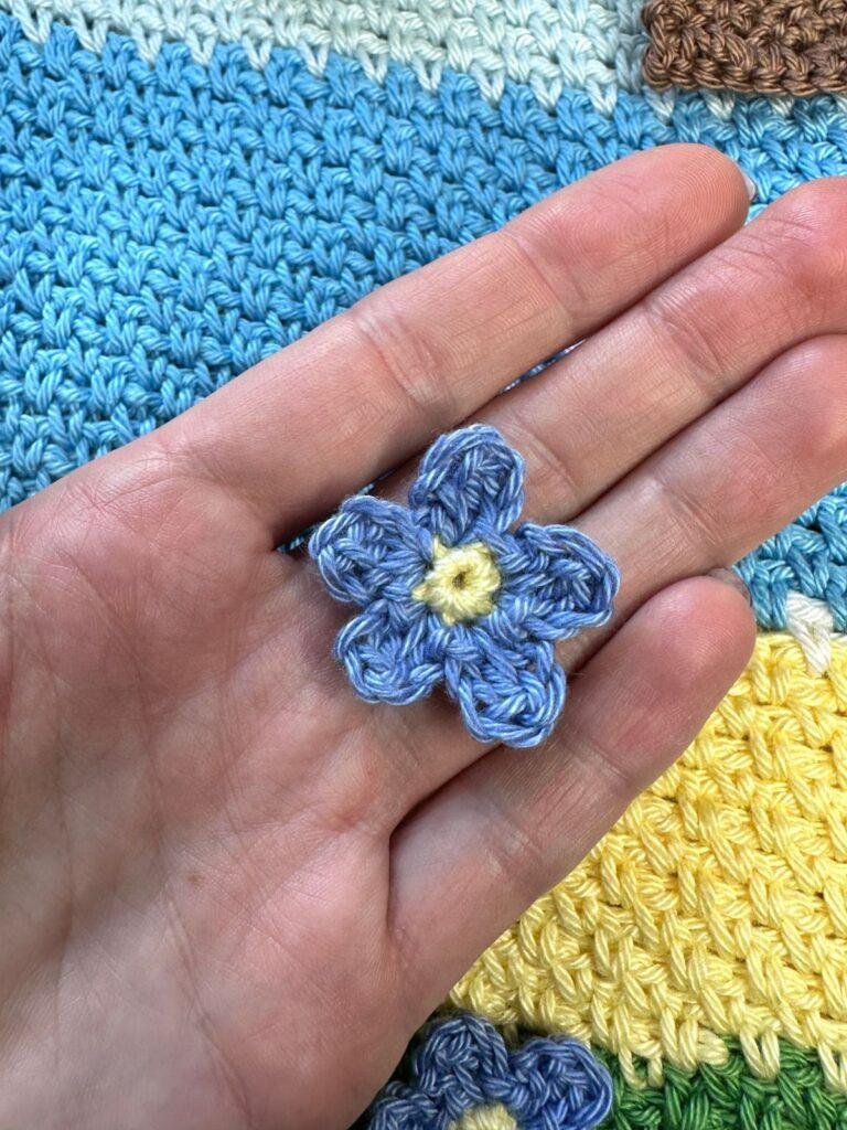 a tiny crochet forget me not flower is held in a hand above a crochet blue and yellow background