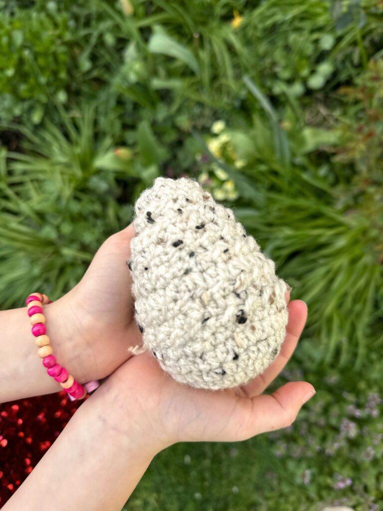 2 small hands hold a tweed rustic crochet Easter Egg over some spring plants