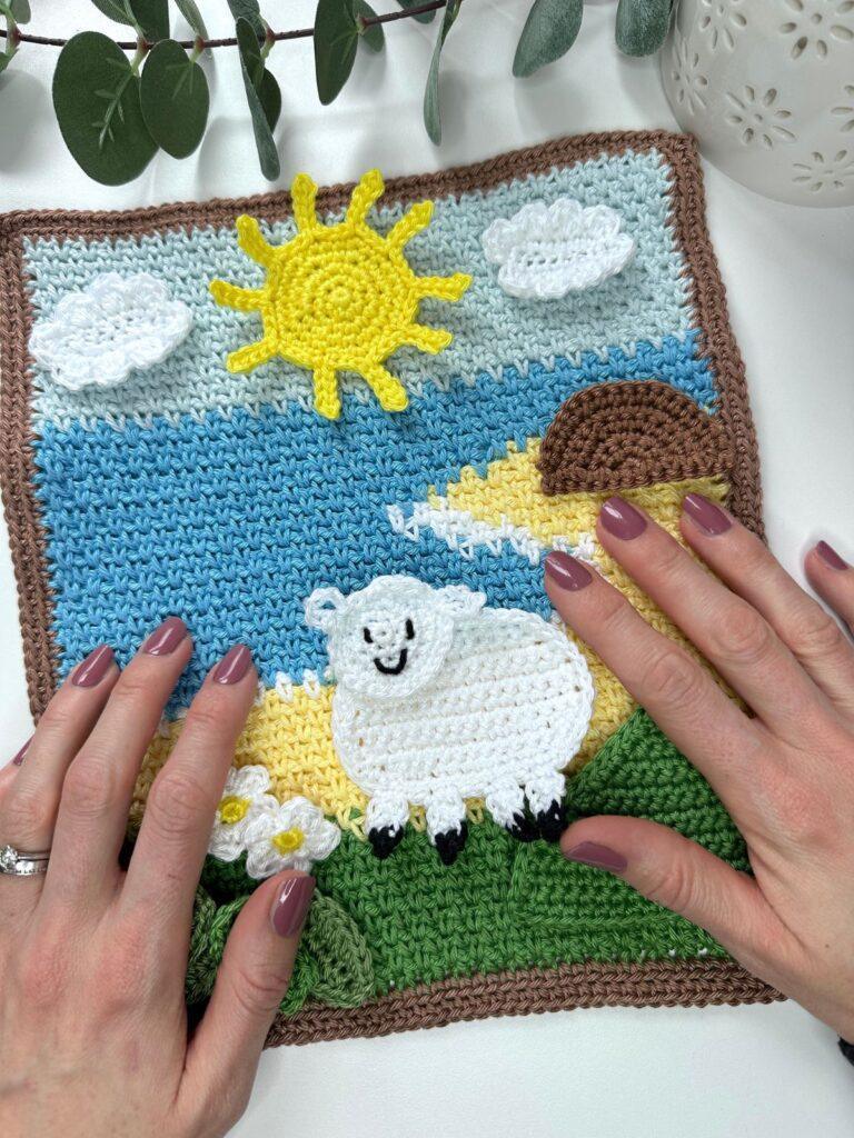 A spring crochet scene of the sea, sun, fields and a sheep have 2 hands over it
