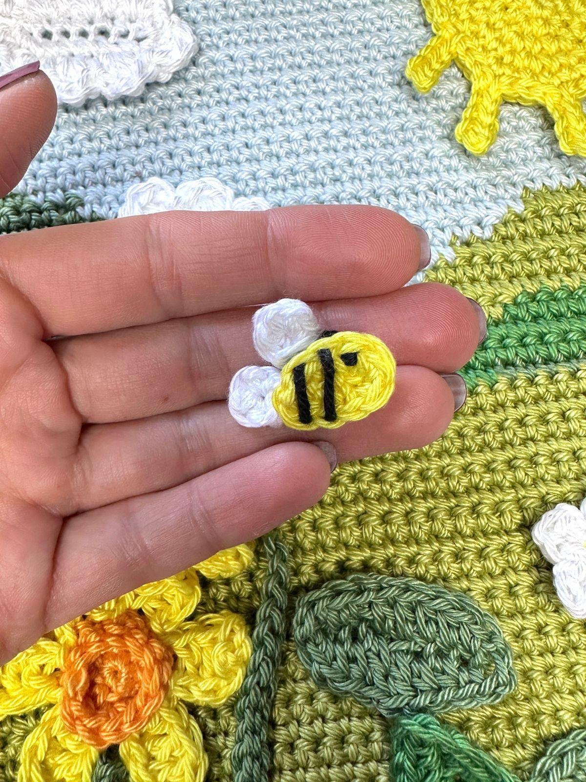 A tiny crochet bee applique is held in a hand above a crochet background