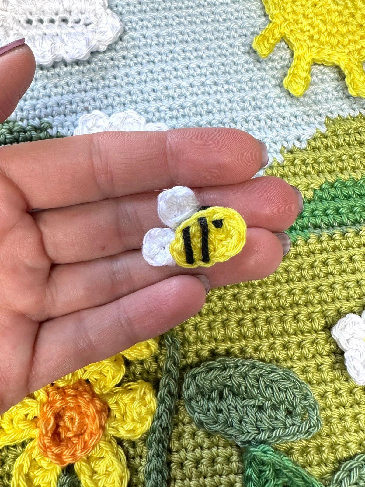 A tiny crochet bee applique is held in a hand above a crochet background