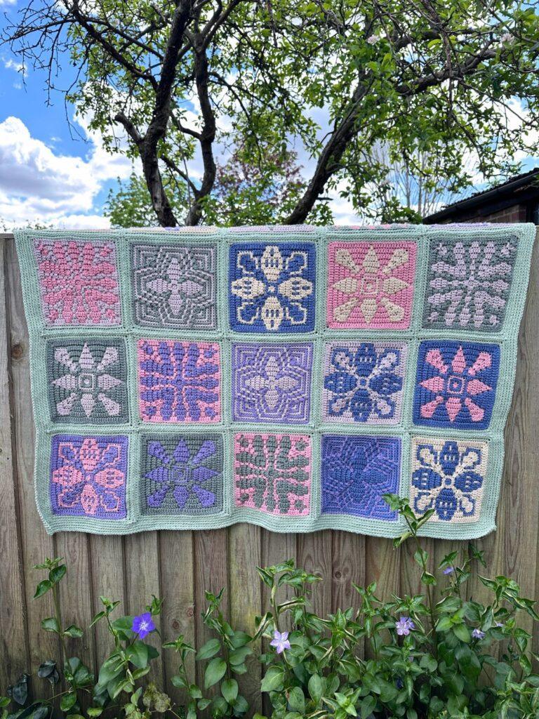 A crochet mosaic blanket of different coloured and patterned squares with a floral theme, is hanging on a fence