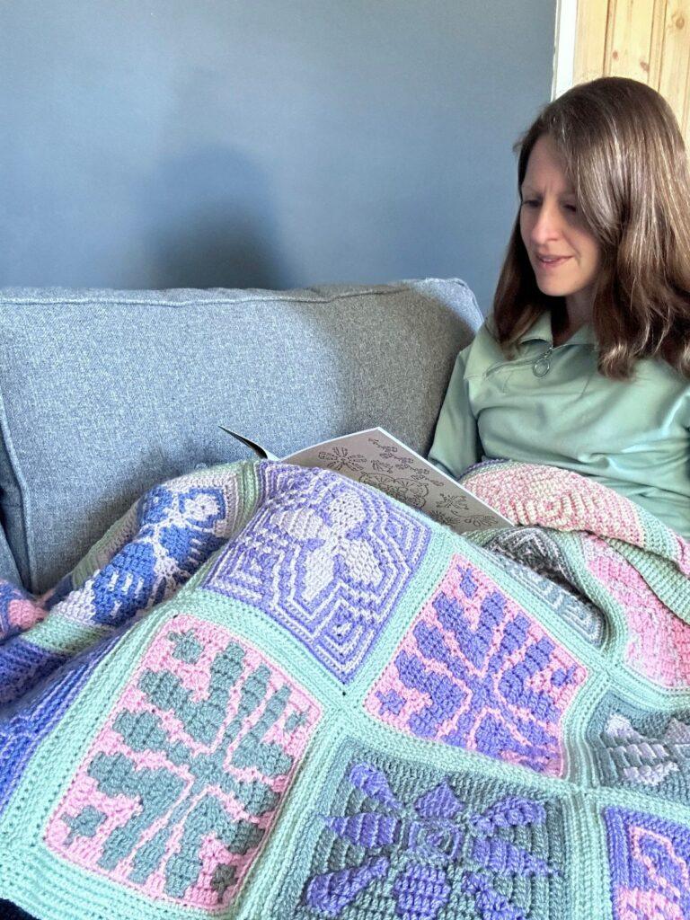 A lady sits under a colourful mosaic crochet blanket reading a book