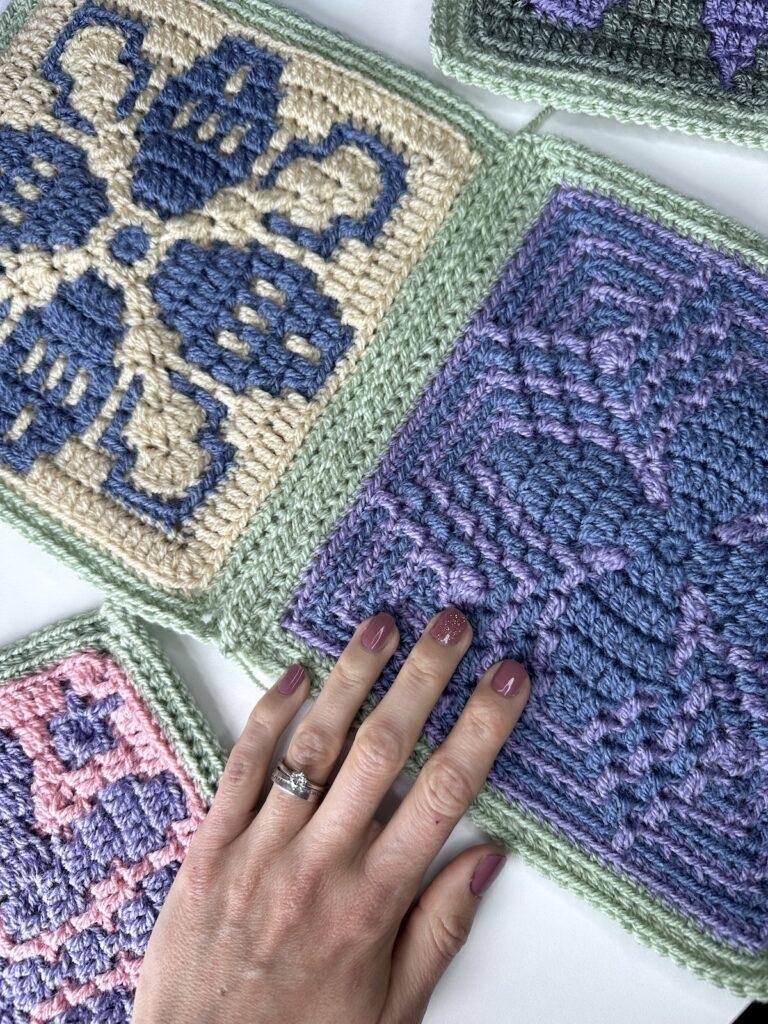 different mosaic crochet squares are on a white surface. 2 squares are joined with a slip stitch seam and a hadn't is touching the square