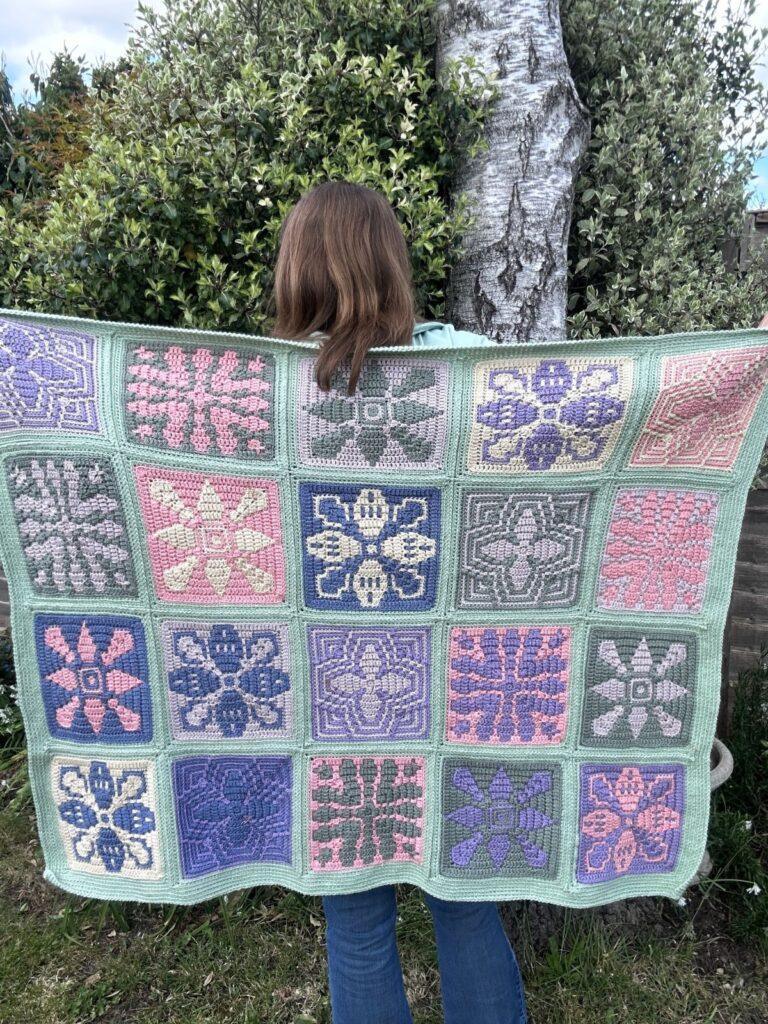 A woman stands in a garden with her back to the camera with a crochet tile blanket held across her back. 