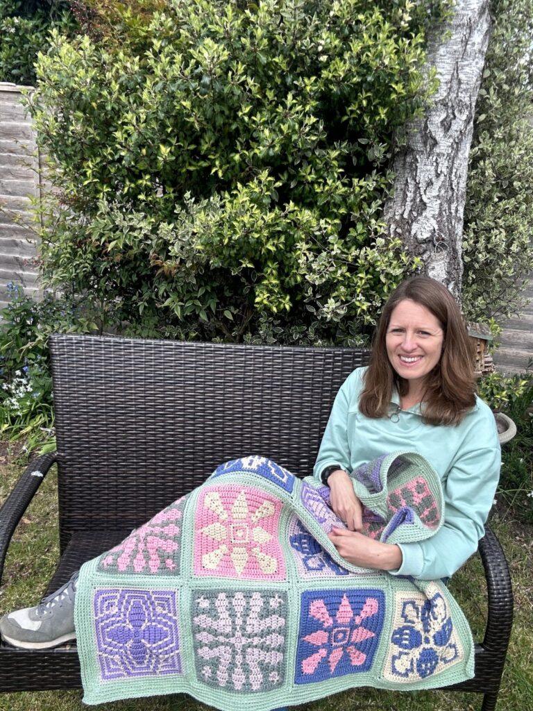 A person sits on a bench by a tree with a mosaic crochet tile blanket over her.