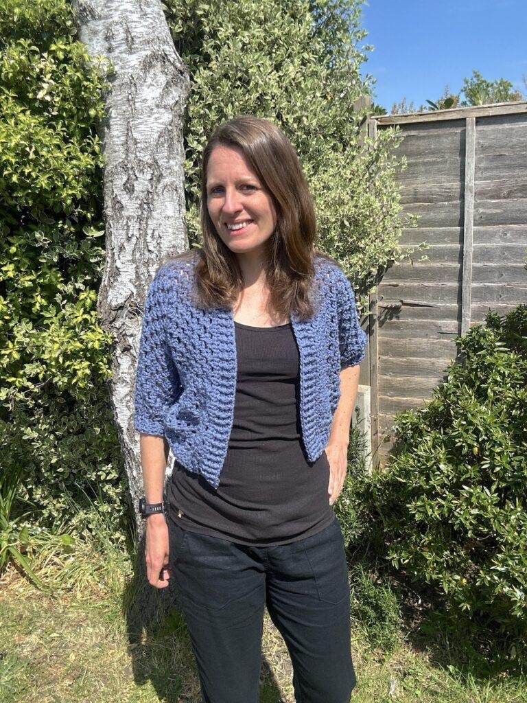 A woman stands in a garden wearing a blue granny stitch style crochet cardigan, black vest and trousers