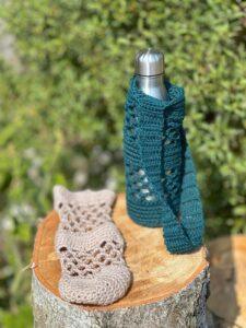 A teal green crochet mesh water bottle holder holds a silver water bottle. Alongside it lies a beige crochet water bottle holder.