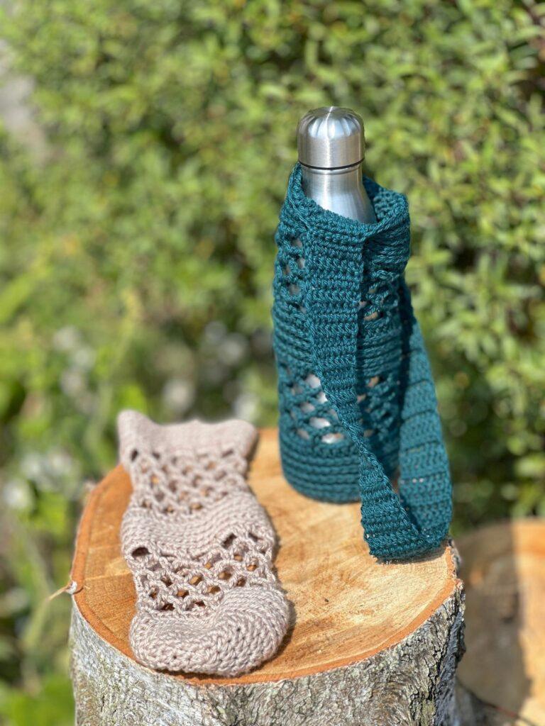 A teal green crochet mesh water bottle holder holds a silver water bottle. Alongside it lies a beige crochet water bottle holder.