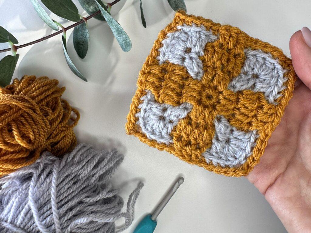 a mosaic crochet center out square in held next to matching orange and grey yarn