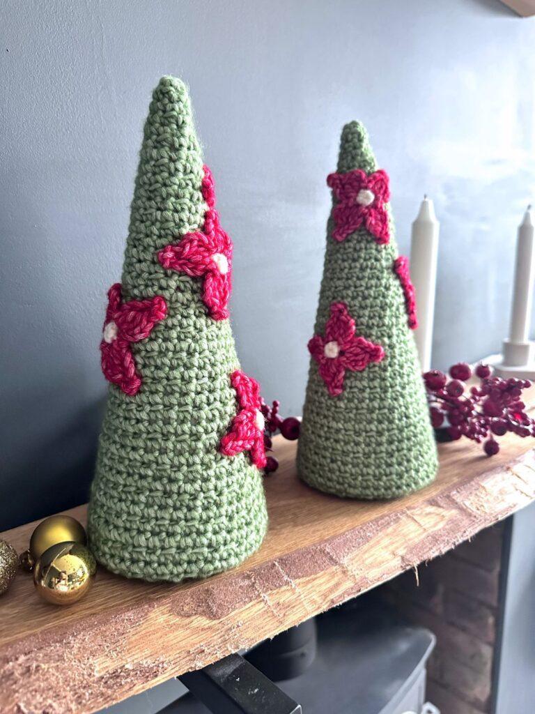 2 crochet Christmas tree cones with flowers on, are stood on a mantelpiece