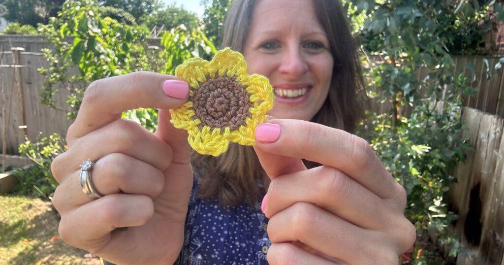 A lady holds a small yellow and brown crochet sunflower motif out in front of her
