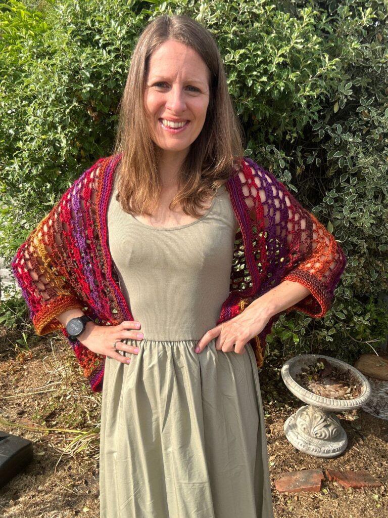 A lady in a green dress wears a crochet cocoon cardigan in red and purple variegated yarn.