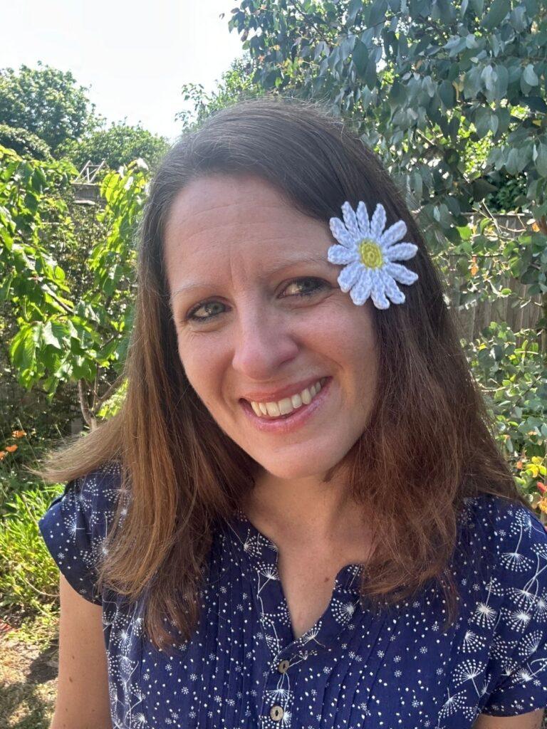 A lady smiling in a garden has a crochet daisy in her hair
